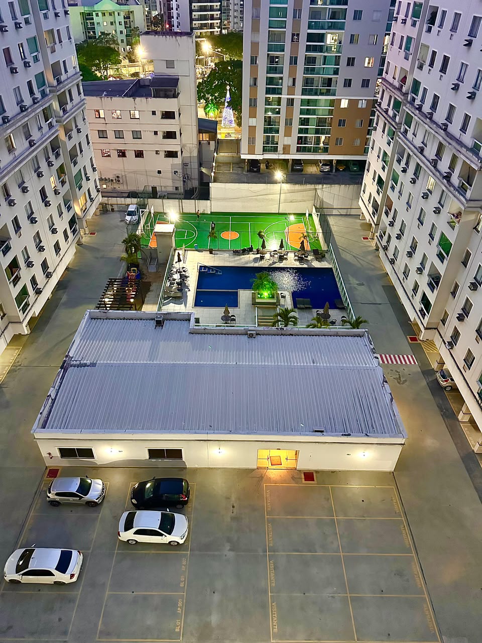 Vista aérea noturna do condomínio mostrando piscina iluminada, quadra e estacionamento